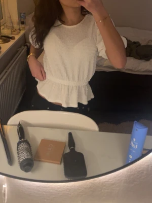 Vit blus med volang och struktur - Supersöt vit blus med volang nertill och lätt puffiga korta ärmar. Toppen har en strukturerad, lite krinklad yta och markerad midja som ger en feminin siluett. Perfekt att styla med jeans eller kjol för en fräsch look.