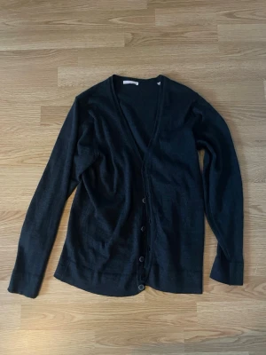 Svart kofta från Knowledge Cotton - Svart, stilren cardigan från Knowledge Cotton Apparel i 100% merinoull. Koftan har v-ringning, långa ärmar och knappar framtill. Perfekt för lager-på-lager och passar dig som gillar minimalistisk design och mjuka material. Nypris: ca 1300