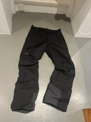 Svarta täckbyxor från Helly Hansen - Svarta täckbyxor från Helly Hansen med regular fit. Byxorna har dragkedjeförsedda fickor och förstärkta partier vid bensluten. Perfekta för vinterns alla äventyr och tillverkade i slitstarkt syntetmaterial.