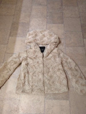 Beige pälsjacka med huva Vero Moda - Säljer en fluffig och mysig beige pälsjacka från Vero Moda med huva. Jackan har långa ärmar och ett snyggt vågigt mönster i pälsen. Insidan är fodrad med slätt tyg och jackan stängs med tryckknappar framtill. Perfekt för kalla dagar!