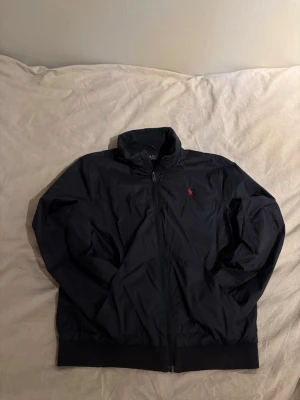 Svart vindjacka från Polo Ralph Lauren - Svart vindjacka från Polo Ralph Lauren med röd logga på bröstet. Jackan har dragkedja framtill, huva som kan tas fram och ribbade muddar vid nederkant. Materialet är lätt och syntetiskt, perfekt för blåsiga dagar. Klassisk och stilren design.