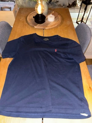 Mörkblå t-shirt från Polo Ralph Lauren - Snygg mörkblå t-shirt från Polo Ralph Lauren med klassisk röd logga broderad på bröstet. Tillverkad i mjuk bomull och har en normal passform med korta ärmar. Perfekt till jeans eller shorts för en clean och stilren look.