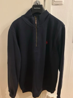 Marinblå half zip tröja Polo Ralph Lauren - Säljer en marinblå långärmad tröja från Polo Ralph Lauren med half zip och röd broderad logga på bröstet. Tröjan har en hög krage och är gjord i mjuk bomull, perfekt för chill dagar. Klassisk och stilren design som funkar till jeans eller chinos. Knappt använd