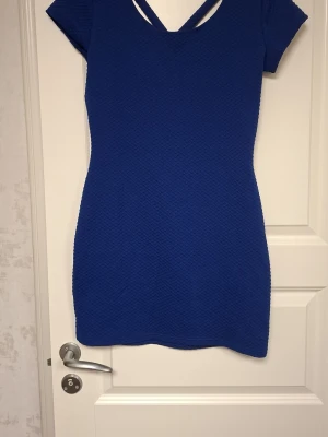 Blå kortklänning med korsade band - Snygg blå kortklänning med strukturmönster och korsade band i ryggen. Klänningen har korta ärmar och en tight passform som framhäver figuren. Materialet är mjukt och stretchigt, perfekt för dig som gillar att sticka ut med färg och detaljer. Tyvärr så har jag klippt bort lappen där det står vart den kommer ifrån.