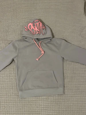 Syna World tracksuit - Säljer denna jättefina Syna World tracksuit eftersom den inte används, knappt använd. Grå tracksuit med Rosa text. Byxorna är i storlek M och hoodien är i storlek S.