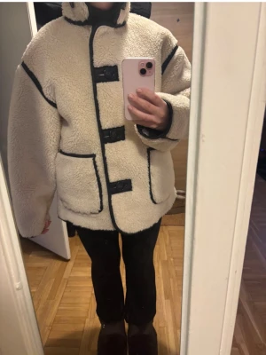Beige teddyjacka med svarta detaljer - Oanvänd. Säljer en beige teddyjacka med svarta kontrastsömmar och stora fickor framtill. Jackan har knäppning med dekorativa spännen och hög krage. Perfekt för dig som vill ha en mysig och trendig look i vinter. Passar både S/M
