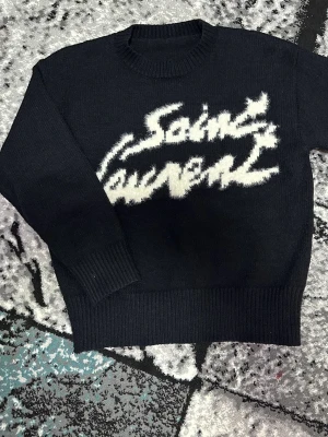 Svart stickad Saint Laurent tröja S - Svart stickad tröja från Saint Laurent med vit fuzzy logotyp framtill. Rund hals, ribbade muddar och nederkant. Tillverkad i Italien, perfekt för dig som gillar exklusiva märken och vill ha en cool statement piece i garderoben.