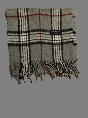 Grå rutig halsduk från Burberrys - Klassisk grå halsduk från Burberrys i 100% lammull med ikoniskt rutigt mönster i svart, vitt och rött. Halsduken har fransar i ändarna och är mjuk och varm – perfekt för kyliga dagar. Perfekt nu när snön har kommit❄️ Kom med frågor eller prisförslag!