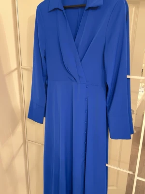 Kornblå långklänning med knappar - Snygg kornblå långklänning med långa ärmar och klassisk krage. Klänningen har omlott-look framtill och dekorativa knappar längs sidan. Materialet känns lätt och följsamt, perfekt för dig som vill ha en stilren och elegant look.