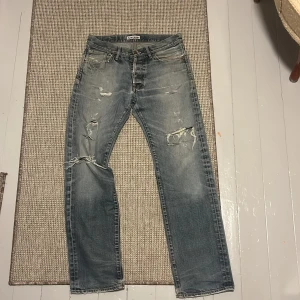 Blå slitna jeans från Acne Jeans - Säljer ett par blå jeans från Acne Jeans med snygga slitningar och hål på benen för en cool, avslappnad look. Jeansen har klassisk femficksdesign, rak passform och är tillverkade i jeansmaterial. Perfekt för dig som gillar streetstyle och vill ha ett par jeans med attityd.