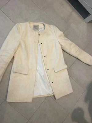 Beige kappa från Zara - Snygg beige kappa från Zara Basic med rund halsringning och knappar framtill. Jackan har två stora fickor och är tillverkad i ullblandning som ger en mjuk och stilren känsla💕Pris kan diskuteras💕