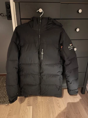 Svart dunjacka Sail Racing - Svart dunjacka från Sail Racing med huva och snygg expedition-patch på ärmen. Jackan är använd 1 vinter då ja växt ut den, inte en ända skada/slitning på den. Kostar 4500kr ny