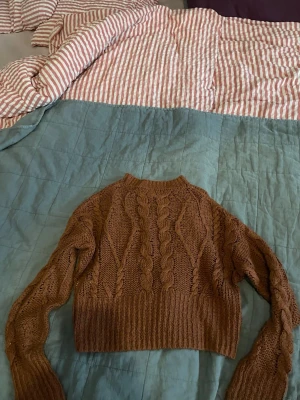 Brun stickad tröja - Säljer en brun stickad tröja med grova flätstickade detaljer och croppad passform. Tröjan har långärmade ärmar och bred ribbad mudd vid midjan och ärmsluten. Perfekt för lager på lager under höst och vinter.