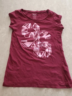 T-shirt - Säljer en vinröd/lila t-shirt från WA Action Sports med coolt grafiskt tryck i ljusrosa på framsidan. T-shirten har rund hals och korta ärmar, perfekt för dig som gillar sportig och avslappnad stil. Materialet är mjuk bomull som känns skönt mot huden. Har ett hål.