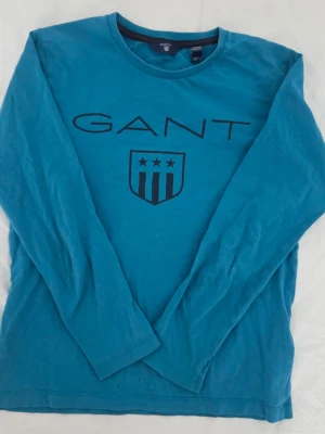 Blå långärmad topp från GANT - Säljer en blå långärmad topp från GANT med tryckt logga och emblem på bröstet. Rund halsringning och enkel design, perfekt för dig som gillar klassiska märken. Materialet är mjuk bomull och toppen har en normal passform.