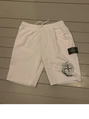 Vita Stone Island shorts med tryck - Säljer ett par vita shorts från Stone Island med elastisk midja och snörning. De har två sidofickor och ett svart logomärke på benet samt ett tryckt kompassmotiv. Perfekta för sommaren och riktigt clean look.