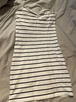 Randig vit tubklänning - Säljer en vit tubklänning med svarta horisontella ränder. Klänningen är axelbandslös och har en enkel, rak passform. Perfekt för sommardagar och enkel att matcha med många skor. Materialet känns mjukt och stretchigt, troligen bomull eller en bomullsblandning.