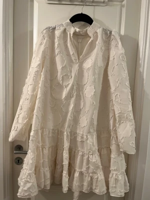 Vit volangklänning från H&M - Säljer en vit klänning från H&M i storlek M. Klänningen har lång ärm, volanger nertill och ett unikt texturmönster med broderade cirklar. Den har en liten ståkrage och är tillverkad i ett lätt, vävt material som ger en luftig känsla.
