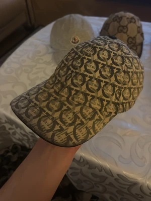 Beige mönstrad keps från Ferragamo - Snygg beige keps från Ferragamo med ett grafiskt mönster i mörkbrunt. Kepsen har justerbar rem baktill och är tillverkad i bomull med mjukt svart innerfoder. Perfekt för dig som gillar statement-accessoarer och vill sticka ut.