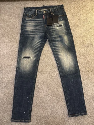 Dsquared2 jeans - Säljer ett par blå jeans från Dsquared2 med coola slitningar och broderade detaljer på bakfickorna. Jeansen har en skinny passform, låg midja och är tillverkade i klassiskt jeanstyg. Färgskiftningar och små färgstänk ger dem en unik look.