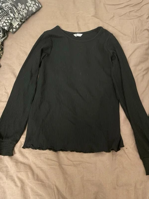 Svart ribbad långärmad topp C - Svart långärmad topp från C i storlek 158/164. Toppen är ribbad med stretchigt material och har smockade muddar vid ärmsluten samt lätt vågig kant nedtill. Enkel och stilren design som funkar till många olika looks.