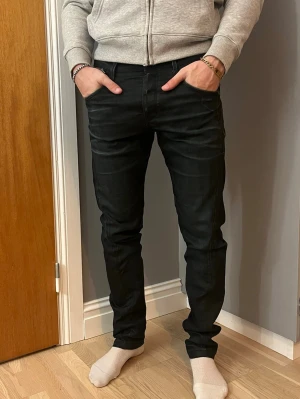 Svarta jeans från Jack & Jones Core - Säljer ett par svarta jeans från Jack & Jones Core med slim passform och klassisk femficksdesign. Byxorna har en snygg dragkedjedetalj vid fickan och raka ben. Materialet är jeans med en mjuk känsla och midwaist. Perfekt för dig som gillar stilrena och moderna jeans.