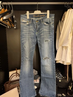 Blå bootcut jeans med slitningar - Snygga blå jeans med bootcut-modell och slitna detaljer på benen samt fransade benslut. Jeansen har klassisk femficksdesign, guldiga broderier på bakfickorna och är tillverkade i jeansmaterial. Perfekta för en avslappnad och trendig look.