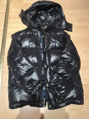 Svart pufferjacka från Polo Ralph Lauren - Säljer en svart pufferjacka från Polo Ralph Lauren med blått innerfoder och broderad blå logga på bröstet. Jackan har huva, dragkedja och tryckknappar samt Polo-detaljer längs dragkedjan. Perfekt för kalla dagar och riktigt snygg streetstil.