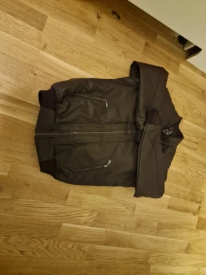 Mörkbrun bomberjacka med dragkedjor - Säljer en mörkbrun bomberjacka från X-Act med ribbade muddar och flera dragkedjeförsedda fickor. Jackan har en klassisk bomberlook med lång ärm och snygga detaljer i metall. Perfekt för dig som vill ha en stilren och enkel jacka.