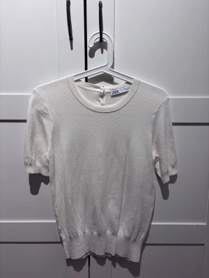 Krämvit kortärmad topp från Zara - Säljer en stilren krämvit topp från Zara med rund halsringning och korta ärmar. Toppen har tre dekorativa knappar bak i nacken och är gjord i ett mjukt, lätt stickat material. Perfekt till jeans eller kjol för en clean look.