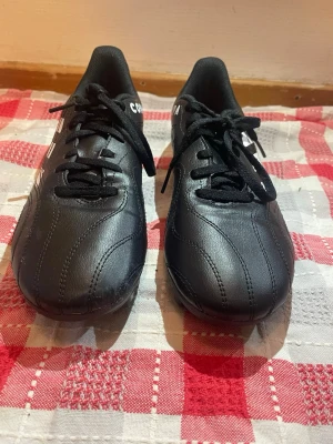 Adidas Copa svarta fotbollsskor - Säljer ett par svarta Adidas Copa fotbollsskor med klassiska vita ränder på sidorna. Skorna har snörning, rund tå och är tillverkade i skinn med syntetdetaljer. Yttersulan har mönster för bra grepp på planen. Perfekta för dig som vill ha stilrena och funktionella skor till matchen.
