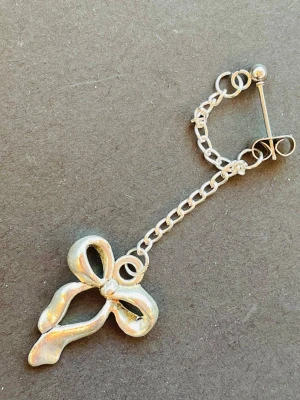 By prisförslag- 1 st Silvrig rosettörhänge med kedja - Unikt örhänge i silverfärgad metall med en kedja och en hängande rosett. Designen är lekfull och detaljrik, perfekt för dig som gillar accessoarer med lite edge. Fästet är klassiskt med plopp och örhänget har en glansig finish som fångar ljuset snyggt.