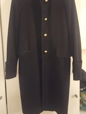 MANGO kappa med guldknappar - Stilren svart kappa med snygga guldknappar framtill och breda manschetter på ärmarna. Klassisk rak modell med två fickor och slät yta. Perfekt för dig som gillar enkel men elegant design och vill ha något som funkar hela hösten och vintern.