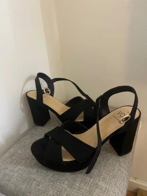 Svarta sandaler med klack från VOX - Ett par svarta sandaler från VOX med breda korsade remmar och justerbart spänne i guld. Skorna har hög blockklack och plattform framtill. Tillverkade i veganskt material med mockakänsla. Har används 1 gång.