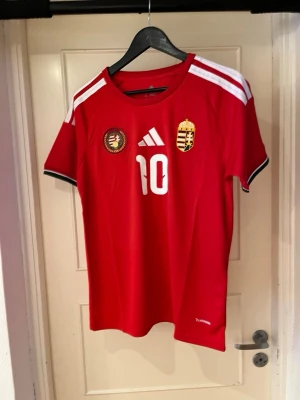 Hungary home kit 25/26 X Szobozslai 10 - Säljer en röd fotbollströja från Adidas med Szoboszlai och nummer 10 på ryggen. Tröjan har vita detaljer, broderat ungerskt landslagsemblem och klassiska Adidas-ränder på axlarna. Materialet är lätt polyester. Om du vill ha eget tryck och ditt namn där bak hör av dig! MVH Moncliqué 