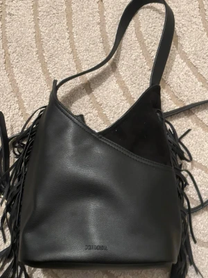Svart axelväska med fransar Don Donna - Säljer en svart axelväska från Don Donna i en mix av skinn och mocka. Väskan har långa fransar på sidorna och en bred axelrem. Snygg asymmetrisk öppning och guldfärgade metalldetaljer. Perfekt storlek för allt du behöver ha med dig. Långa bandet tillkommer😃 Köpt 899kr