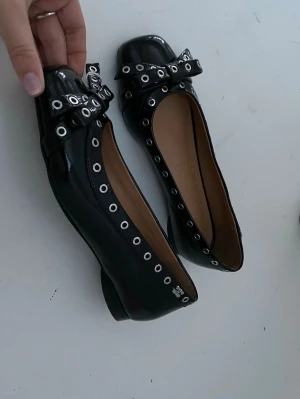 Svarta ballerinaskor med rosett från Ganni - Oanvända svarta ballerinaskor från Ganni i lackat skinn med silverfärgade nitar runt kanten och en cool rosett framtill. Skorna har fyrkantig tå och låg klack, levereras med originalkartong och dustbag. Perfekt för dig som gillar edgy detaljer och stilren design. 