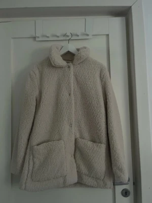 Beige teddyjacka från H&M - Mysig beige teddyjacka från H&M med stora framfickor och hög krage. Jackan är långärmad och har en rak passform, perfekt för kyliga dagar. Stängs med knappar framtill och har en fluffig, mjuk känsla. 