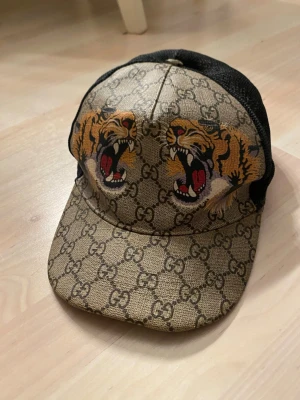 keps med tigertryck - Säljer en snygg  keps med klassiskt GG-mönster i beige och svart mesh på sidorna. Framtill är det två coola tigerhuvuden med öppna käftar i orange, svart och rött. Justerbar baktill med grönt och rött band. Perfekt för dig som vill sticka ut.