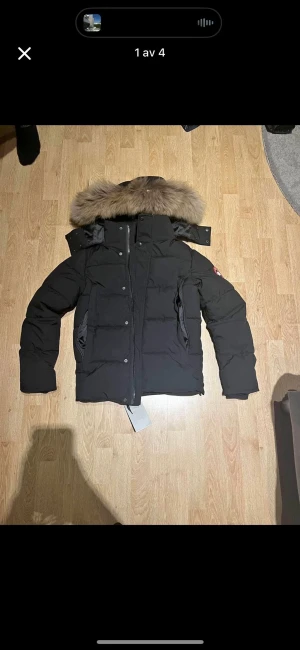 Svart dunjacka från Canada Goose - Svart dunjacka från Canada Goose med fluffig beige päls på huvan. Jackan har quiltad design, dragkedja och knappar framtill samt långärmade ärmar. Klassisk patch på ärmen och flera fickor. Perfekt för kalla vinterdagar.