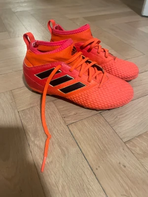 Röda adidas fotbollsskor  - Snygga och bekväma fotbollsskor från Adidas i stark orange färg. Kardborre/strumpliknande skaft som ger bra stöd runt vristen och snörning för optimal passform. Passar perfekt för träning och match på konstgräs/gräs.