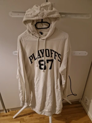 Vit hoodie PLAYOFFS 87 H&M S - Vit hoodie från H&M med svart tryck 'PLAYOFFS 87' på bröstet. Klassisk modell med huva och dragsnören, samt känguruficka framtill. Mjuk insida och långa ärmar. Perfekt för en sportig och avslappnad look.