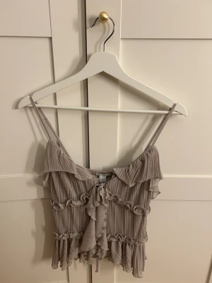 Beige volangtop från Amisu - Superfin beige linnetopp från Amisu med tunna axelband, volangdetaljer och snörning framtill. Toppen har ribbad struktur och är croppad med romantiska ryschkanter. Perfekt för dig som gillar en söt och trendig look.