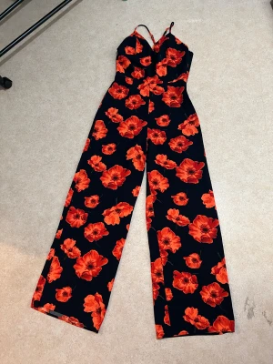 Svart byxdress med röda blommor - Säljer en snygg svart byxdress med breda ben och tunna axelband. Plagget har ett iögonfallande blommigt mönster i orangerött, samt en v-ringad överdel. Perfekt för dig som vill sticka ut med färg och mönster. Materialet känns mjukt och följsamt. Storlek XS, men passar även S (ca 34–36), själv hade s/M i storlek. Använts max 4 gånger, i jättebra skick – inga defekter.