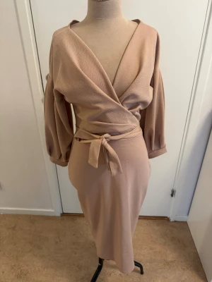 Beige omlottklänning från Boohoo - Snygg beige omlottklänning från Boohoo med knyt i midjan och v-ringad hals. Klänningen har långa, puffiga ärmar och en figurnära passform som ger en elegant siluett. Perfekt för dig som gillar stilrena och trendiga plagg.