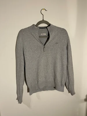 Grå GANT half zip tröja - Snygg grå långärmad tröja från GANT med half zip och broderad logga på bröstet. Tröjan har ribbad krage och muddar, tillverkad i mjuk bomull som är skön mot huden. Har ett litet hål i ena armhålan men är fixat. Säljs såklart för lägre pris pga det. Köp på NK. Storlek 170 cm.