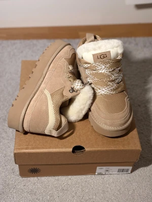 Beige UGGlowmel  - UGG boots i beige mocka och textil med grov sula och fluffigt vitt fårullsfoder. Skorna har breda snören i beige och vitt, UGG-logga på plös och häl, samt robust yttersula för extra grepp. Perfekta för kalla dagar och snygg streetstil.