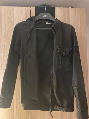 Svart overshirt från Stone Island - Svart overshirt från Stone Island med klassisk patch på ärmen. Jackan har dragkedja framtill, bröstficka med knapp och lång ärm. Det är bara att skriva vid minsta fundering. Storlek S men passar M kan gå ner i pris vid snabb affär