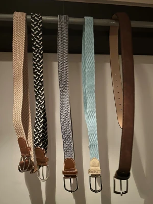 Fem snygga bälten i olika färger - Säljer fem bälten i olika färger och mönster. Ett beige flätat bälte, ett svartvitt randigt, ett gråvitt randigt, ett ljusblått flätat och ett brunt mockabälte. Alla har metallspännen och detaljer i helgen skinn eller syntet. Perfekt för att lyfta din outfit.