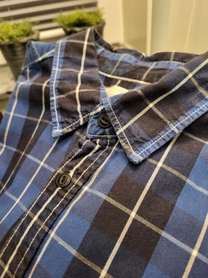 Vintage Blårutig skjorta från Levi's - Snygg blårutig skjorta från Levi's i bomull. Skjortan har klassisk krage, svarta knappar och långa ärmar. Mönstret är rutor i blått, svart och vitt. Perfekt för dig som gillar en avslappnad men stilren look.